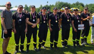 Grupa siedmiu strażaków w czarnych mundurach galowych z medalami na szyjach oraz Zastępca Burmistrza Drzewicy w cywilnym ubraniu pozują do zdjęcia na trawiastym boisku w słoneczny dzień. Jeden ze strażaków trzyma czek okolicznościowy na kwotę 2.000 zł, a inny puchar. W tle widoczne są namioty i drzewa.
