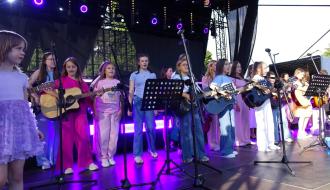 Występ dziecięcego zespołu muzycznego na dużej scenie. Dziewczynki śpiewają i grają na gitarach akustycznych podświetlone fioletowymi reflektorami.