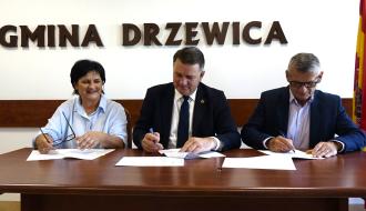 Przy długim, drewnianym stole konferencyjnym siedzą – od lewej: w jasnej koszuli radna Rady Powiatu w Opocznie Teresa Szklarczyk trzymająca okulary, Członek Zarządu Woj. Łódzkiego Artur Bagieński i Burmistrz Drzewicy Janusz Reszelewski. Obaj w garniturach składają podpisy na leżących przed nimi dokumentach. W tle duży napis na ścianie „GMINA DRZEWICA”, nad nim godło Polski oraz herb miejscowości. Po prawej stronie flaga.