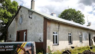 Stary, parterowy budynek „Ceglanki” ze spękaną elewacją. Przed nim stoi oklejona furgonetka z napisem informującym o robotach ziemnych i wykopach.