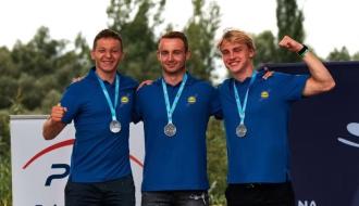 Trzej uśmiechnięci kajakarze górscy w niebieskich koszulkach polo z logo LKK Drzewica – od lewej: Szymon Zawadzki, Michał Franczak, Maciej Bartos - stoją obok siebie na podium, obejmując się ramionami. Każdy z nich ma na szyi srebrny medal.  W tle banery sponsorskie oraz zieleń drzew.