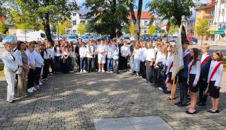 Zgromadzona w parku na Placu Wolności młodzież szkolna i nauczyciele SP w Drzewicy obserwują uroczystość. Po prawej stronie stoi poczet sztandarowy.
