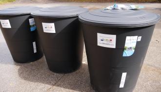 Trzy duże, czarne plastikowe zbiorniki na deszczówkę stojące w rzędzie na asfalcie. Na każdym z nich naklejona jest tabliczka informacyjna. 