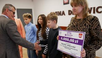  Burmistrz Drzewicy w marynarce i okularach uściska dłoń chłopca trzymającego symboliczny czek finansowy programu „Kreatywna Młodzież 2026” na kwotę 6 000,00 zł. Obok stoją inni uczniowie oraz nauczycielka w sukience we wzór. W tle widnieje godło Polski, herb oraz napis na ścianie „Gmina Drzewica”. 