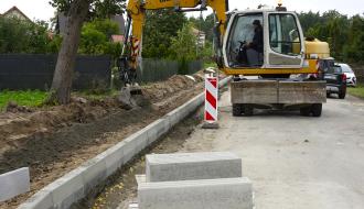 Żółta koparka kołowa marki Liebherr pracująca przy wykopie pod krawężnik na placu budowy drogi powiatowej Drzewica - Dąbrówka. Na pierwszym planie leżą betonowe krawężniki na palecie.