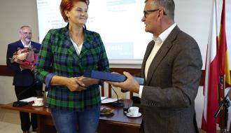 Przewodnicząca Towarzystwa Przyjaciół Drzewicy w zielono-niebieskiej żakietowej marynarce w kratę i dżinsach odbiera podłużne ozdobne etui od Burmistrza Drzewicy w okularach i marynarce. W tle po lewej stronie stoi Wiceprzewodniczący Rady Miejskiej w garniturze, trzymający bukiet kwiatów. Na tylnej ścianie ekran projekcyjny.