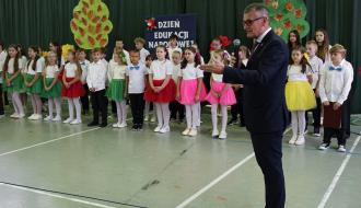 Burmistrz Drzewicy w ciemnym garniturze i okularach przemawia do mikrofonu na hali sportowej. W tle stoi grupa dzieci w galowych strojach z kolorowymi spódniczkami i muszkami. W tle widnieje tablica z napisem „Dzień Edukacji Narodowej”. 