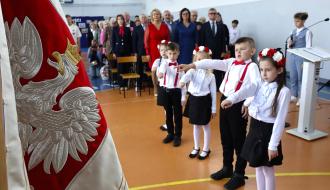 Dzieci w galowych strojach z wiankami i czerwonymi szelkami składają ślubowanie z podniesioną prawą ręką podczas uroczystości w sali gimnastycznej. Na pierwszym planie fragment sztandaru z godłem Polski. W tle stoją goście i nauczyciele. 