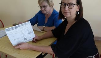 Dwie kobiety siedzą przy stole. Jedna patrzy w obiektyw i trzyma otwartą broszurę, druga patrzy na stół.