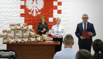 Uroczyste spotkanie w sali USC. Przed stołem uginającym się pod pluszowymi misiami i upominkami stoi Burmistrz Drzewicy w garniturze z czerwoną teczką oraz dwie kobiety – Dyrektor CUS i pracownica CUS. W tle wielkie godło Polski na biało-czerwonym tle. 