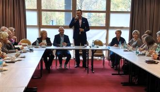 Burmistrz Nałęczowa w garniturze przemawia do mikrofonu, stojąc na środku sali przed rzędem okien, podczas spotkania z grupą osób siedzących przy stołach. 