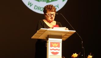 Pani Prezes UTW w okularach i czarnej sukience przemawia przy mównicy ozdobionej herbem Drzewicy. W tle widoczne jest logo Uniwersytetu Trzeciego Wieku w Drzewicy. 