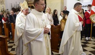 Procesja wejścia w drzewickim kościele. Duchowni w białych szatach liturgicznych, na czele z Arcybiskupem Depo w mitrze, idą nawą główną wzdłuż ławek wypełnionych wiernymi. W tle widoczny jest ołtarz oraz osoby trzymające sztandar.