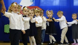 Dzieci ubrane na galowo, trzymając się za ramiona, idą w rzędzie w sali przedszkolnej podczas uroczystości pasowania.