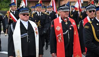 Uroczysta parada strażacka. Na pierwszym planie ks. Grzegorz Włodarczyk i ks. Leszek Sokół w czarnych mundurach strażackich. Duchowny po lewej ma założoną białą stułę, a duchowny po prawej czerwoną stułę kapelańską. Za nimi ulicą maszeruje kolumna strażaków OSP w galowych mundurach oraz poczty sztandarowe z flagami. 
