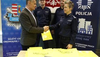 Burmistrz Drzewicy Janusz Reszelewski przekazuje kamizelkę odblaskową na ręce Komendanta Powiatowego Policji w Opocznie, mł. insp. Magdaleny Kucharskiej. Z tyłu dwaj funkcjonariusze Policji