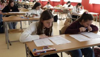 Uczniowie siedzący w ławkach w sali lekcyjnej biorą udział w pisemnej części konkursu, rozwiązując test