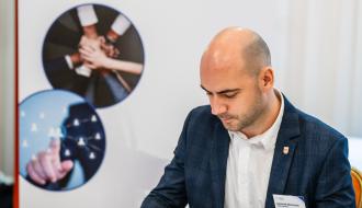 Zastępca Burmistrza Drzewicy Dominik Niemirski podpisuje list intencyjny, siedząc przy stole z białym obrusem. W tle roll-up Łódzkiej Akademii Wsparcia Małego Dziecka