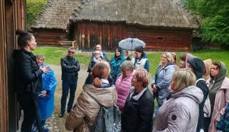 Kilkunastoosobowa grupa z Gminy Drzewica, stojąc na tle drewnianych, krytych strzechą chat, słucha pani przewodnik w Muzeum Wsi Lubelskiej
