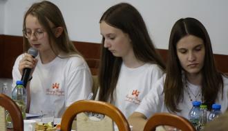 Trzyosobowy zespół uczennic SP w Domasznie składa sprawozdanie z realizacji projektu grantowego. Uczennice siedzą przy stole. Jedna zabiera głos, trzymając mikrofon
