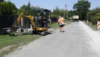 Roboty na poboczu remontowanej drogi. Po lewej pracująca minikooparka oraz pracownik drogowy