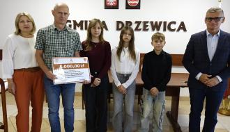  Na tle ściany z napisem GMINA DRZEWICA, godłem Polski i herbem Drzewicy od lewej stoją: Dyrektor ZEAS Małgorzata Kuchenbecker, nauczyciel (trzyma plastikową promesę) i troje uczniów SP w Idzikowicach, Burmistrz Drzewicy Janusz Reszelewski.