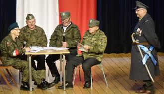 Występ podopiecznych Dziennego Domu Senior+ w Drzewicy. Na scenie pięć osób ubranych w stroje wojskowe. Jedna pani i trzech panów siedzi przy stole. Obok po prawej stoi pan grający Marszałka Józefa Piłsudskiego