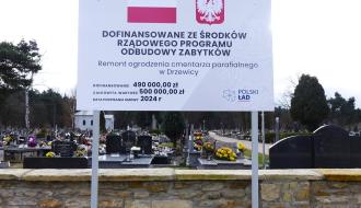 Stojąca przed murem tablica informująca o dofinansowaniu inwestycji ze środków Rządowego Programu Odbudowy Zabytków