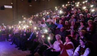 Słuchacze zgromadzeni na widowni trzymają w dłoniach telefony z zapalonymi latarkami