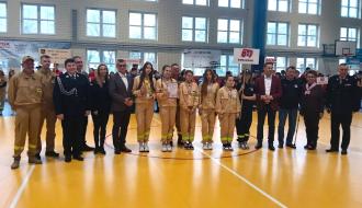 Mistrzowska drużyna dziewcząt z OSP Domaszno prezentuje zdobyte trofea, stojąc w hali sportowej w otoczeniu organizatorów i gości zawodów.