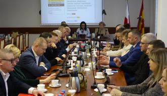 Radni i inni uczestnicy sesji siedzą przy stołach w sali konferencyjnej Urzędu Miejskiego w Drzewicy. Z tyłu na środku stół prezydialny i ekran.