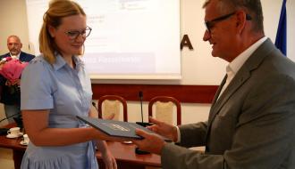 Burmistrz Drzewicy w szarym garniturze i okularach wręcza ciemnoniebieską teczkę z godłem Pani Dyrektor Przedszkola Samorządowego w Drzewicy w błękitnej sukience.