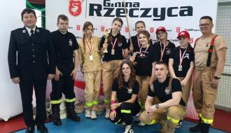 Drużyna dziewcząt z OSP Domaszno wraz z opiekunami oraz Gminnym Komendantem ZOSP w Drzewicy stoi na tle baneru z napisem Gmina Rzeczyca. Dziewczęta mają medale i puchar.