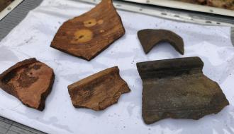  Pięć fragmentów średniowiecznych naczyń ceramicznych ułożonych na zafoliowanej białej kartce papieru.