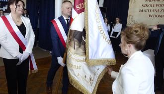 nętrze szkoły podczas uroczystości. Mężczyzna w garniturze z biało-czerwoną szarfą trzyma sztandar szkoły w kolorze biało-czerwonym z haftem. Naprzeciw kobieta w jasnym stroju, dotykająca dolnej części sztandaru. Obok kobieta z pocztu sztandarowego, również z biało-czerwoną szarfą. 