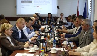 Posiedzenie przy długim stole konferencyjnym. Radni i inni uczestnicy sesji w formalnych strojach siedzą po obu stronach, korzystają z tabletów i dokumentów. W tle widoczny ekran z wynikami głosowania oraz flagi. Na stole znajdują się mikrofony, kawa i napoje.