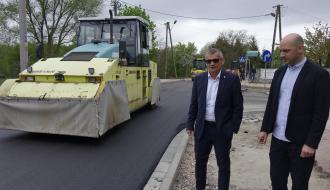 Po prawej stoją Burmistrz i Zastępca Burmistrza Drzewicy, którzy oglądają prace drogowe na ulicy Braci Kobylańskich. Po lewej na jezdni walec drogowy zagęszczający świeży asfalt. W tle kolejny walec drogowy. Niebo zachmurzone.