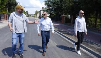 Trzech mężczyzn idzie środkiem nowo wyasfaltowanej drogi i rozmawia. Mężczyzna po lewej, w szarej bluzie, żywo gestykuluje. W środku mężczyzna w okularach przeciwsłonecznych, jasnej koszuli i jeansach, a po prawej mężczyzna w jasnej koszuli w paski i czarnych spodniach. W tle domy jednorodzinne i drzewa.