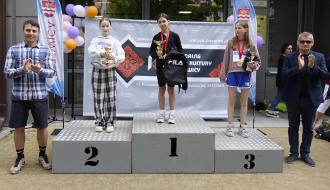 Troje dziewcząt z pucharami i medalami na podium przed RCK w Drzewicy. Laureatka na pierwszym miejscu trzyma czarny plecak. Po bokach asystują Wiceprezes LGD „Nad Drzewiczką” i Burmistrz Drzewicy, klaszcząc w dłonie. W tle ścianka reklamowa i balony.
