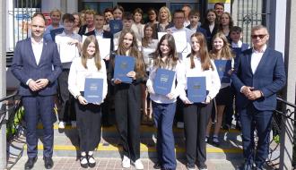 Oficjalne zdjęcie grupowe nowo wybranych radnych Młodzieżowej Rady Miejskiej w Drzewicy wraz z dorosłymi, w tym Burmistrzem Januszem Reszelewskim, na schodach przed budynkiem urzędu. Młodzież trzyma niebieskie teczki z herbem gminy.