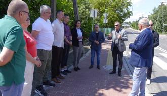 Grupa urzędników i radnych rozmawia na nowym czerwonym chodniku przy drodze. Tyłem stoi I Wicewojewoda Łódzki w niebieskiej marynarce, który żywiołowo gestykuluje.