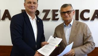Członek Zarządu Woj. Łódzkiego Artur Bagieński i Burmistrz Drzewicy Janusz Reszelewski stoją obok siebie i podają sobie dłonie w geście uścisku, trzymając w rękach podpisane dokumenty. W tle widoczny jest fragment napisu na ścianie „GMINA DRZEWICA”.