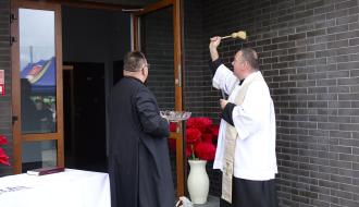 Uroczystość poświęcenia budynku. Dwóch duchownych w szatach liturgicznych stoi przy otwartych drzwiach wejściowych. Jeden z nich unosi kropidło, święcąc obiekt wodą, podczas gdy drugi trzyma naczynie. Obok wejścia stoi stół nakryty białym obrusem oraz wazon z dużymi, sztucznymi makami.
