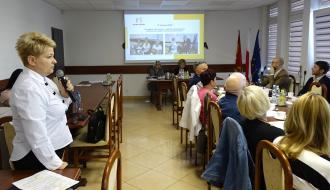 Kobieta z krótkimi blond włosami przemawia do mikrofonu, stojąc po lewej stronie sali konferencyjnej. W głębi sali, na ścianie, wyświetlana jest prezentacja multimedialna, a po prawej stronie, przy stole, tyłem do obiektywu siedzą uczestnicy spotkania.