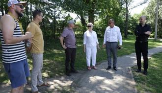 Grupa sześciu osób w letnich i eleganckich strojach rozmawia na parkowej ścieżce obok placu zabaw