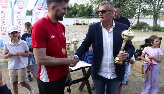 Burmistrz Drzewicy w garniturze wręcza puchar za trzecie miejsce i podaje rękę siatkarzowi w czerwonej koszulce z medalem na szyi. Z tyłu dzieci i flagi promocyjne Drzewicy.