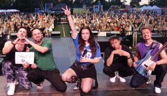 Zespół muzyczny z Lanberry w środku pozuje na scenie w kuckach, tyłem do licznie zgromadzonej publiczności. Jeden z muzyków trzyma kartkę z napisem PAWCIO.