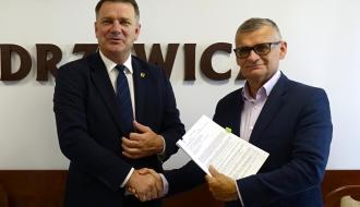 Członek Zarządu Woj. Łódzkiego Artur Bagieński i Burmistrz Drzewicy Janusz Reszelewski ściskają sobie dłonie na tle ściany z częściowo zakrytym napisem „DRZEWICA” Burmistrz trzyma podpisany dokument.