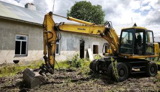 Żółta koparka kołowa Komatsu zaparkowana na rozkopanym, porośniętym chwastami podwórzu przed spękaną elewacją budynku „Ceglanka”.