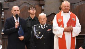 Cztery osoby stojące w kościele podczas uroczystości. Po lewej stronie Wiceburmistrz Drzewicy w ciemnym płaszczu przemawia do mikrofonu, trzymając teczkę. Pośrodku stoi starszy mężczyzna w galowym mundurze strażackim z odznaczeniami. Po prawej stronie stoi ks. Leszek Sokół w białej albie i czerwonej stule, trzymający w dłoniach czerwoną księgę. 